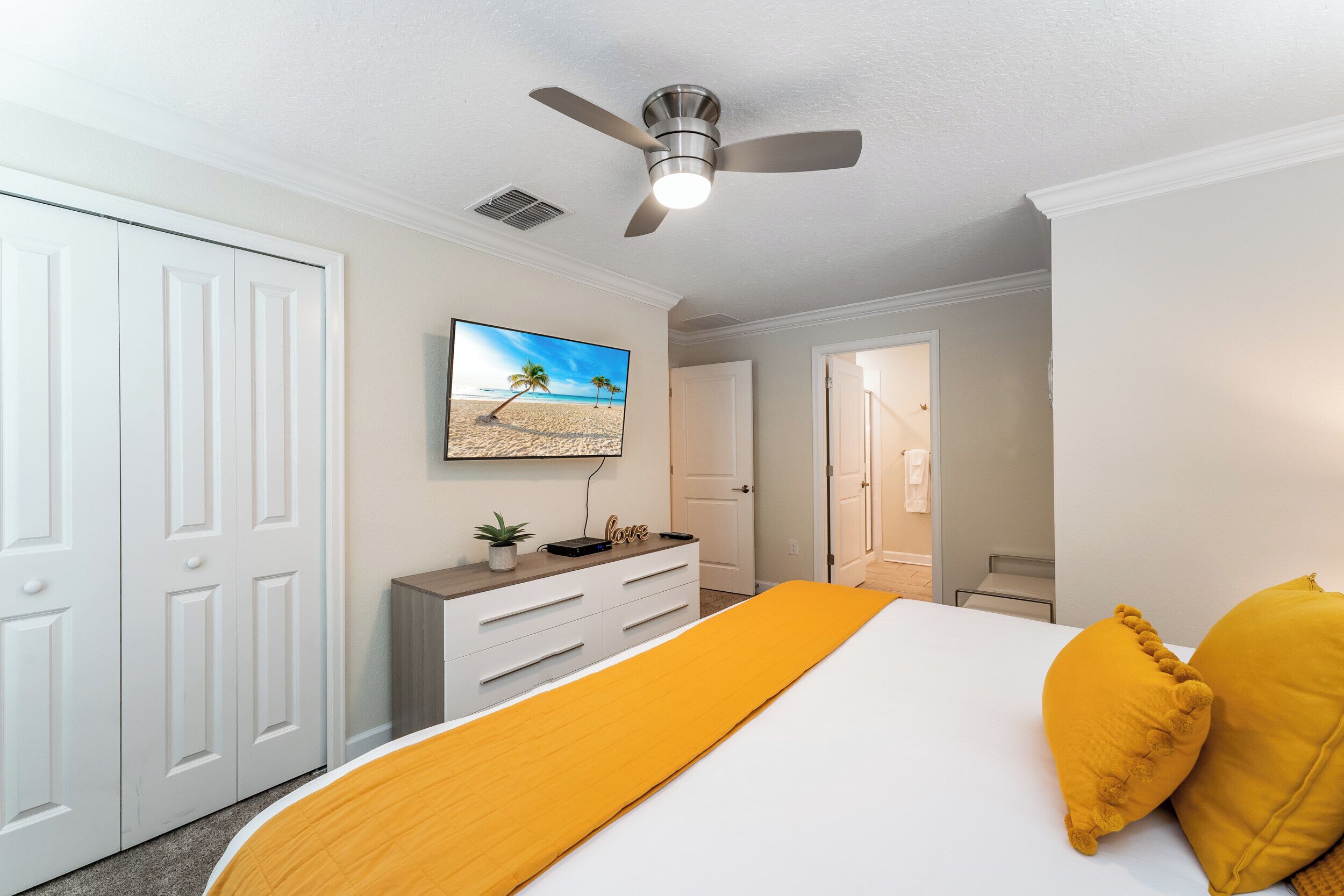 A bedroom with a bed, a ceiling fan, a TV, a dresser, and a door leading to another room.