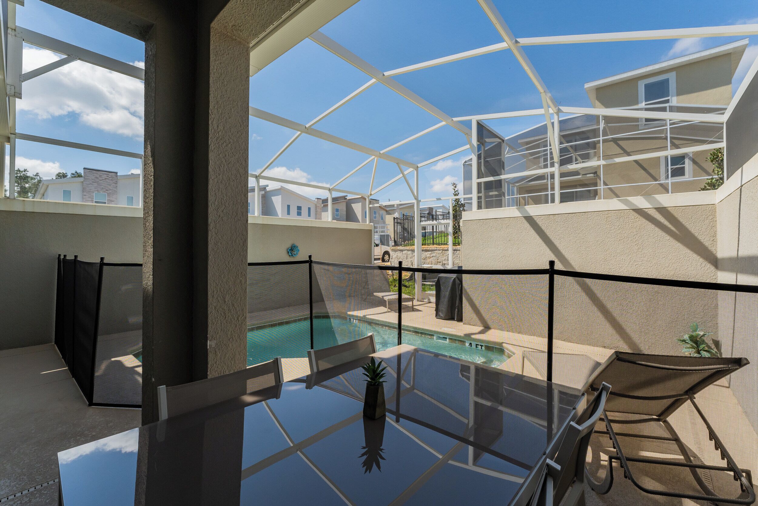A glass-enclosed pool area with lounge chairs and a small table, surrounded by a white fence.