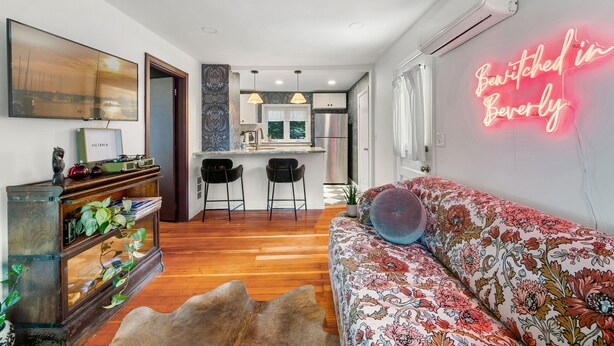 Uma sala de estar moderna com sofá, mesa de centro e televisão em um suporte. Cozinha visível ao fundo, com geladeira e micro-ondas.