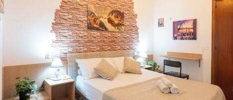 A bedroom with a brick wall, a bed with a beige bedspread, a nightstand with a lamp, a chair, and a small table with a plant.