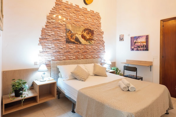 A bedroom with a brick wall, a bed with a beige bedspread, a nightstand with a lamp, a chair, and a small table with a plant.