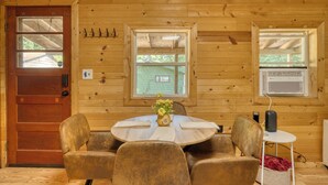 A wooden cabin interior with a round dining table, four chairs, a small side table, and a door with a window. - Cozy Log Cabin with Dock, Kayaks, Firepit, AC, minutes to Ludington and Lake MI (Fountain)