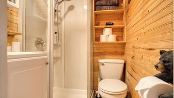 A wooden cabin bathroom with a glass shower enclosure, a toilet, and a shelf with towels and toiletries.