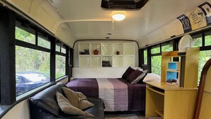 Un interior compacto y bien iluminado con una cama, un sofá de cuero negro y un pequeño escritorio de madera. Amplias ventanas dejan entrar luz natural y ofrecen vistas al exterior.