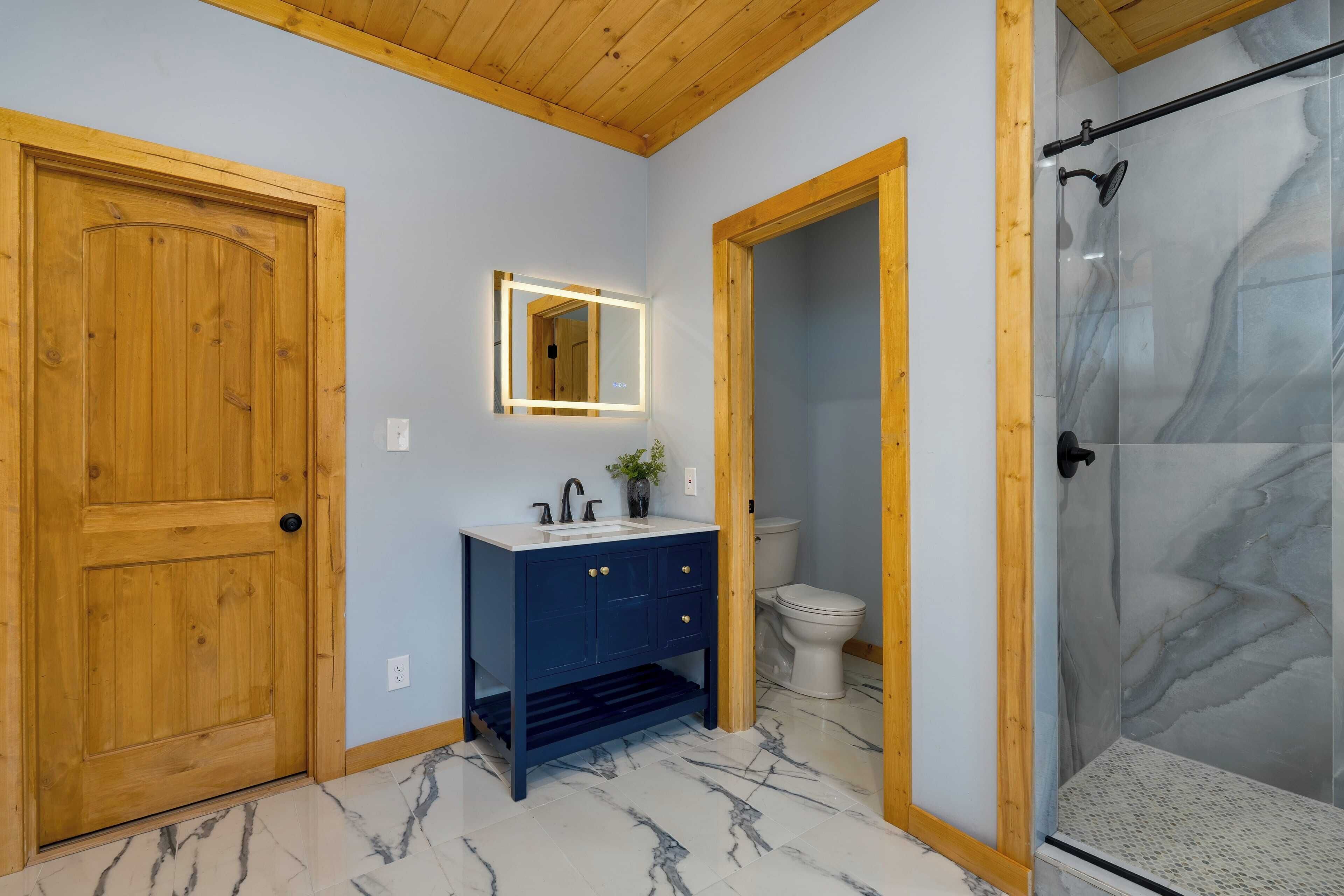 A bathroom with a wooden door, a dark blue vanity with a mirror, a toilet, and a glass shower enclosure.