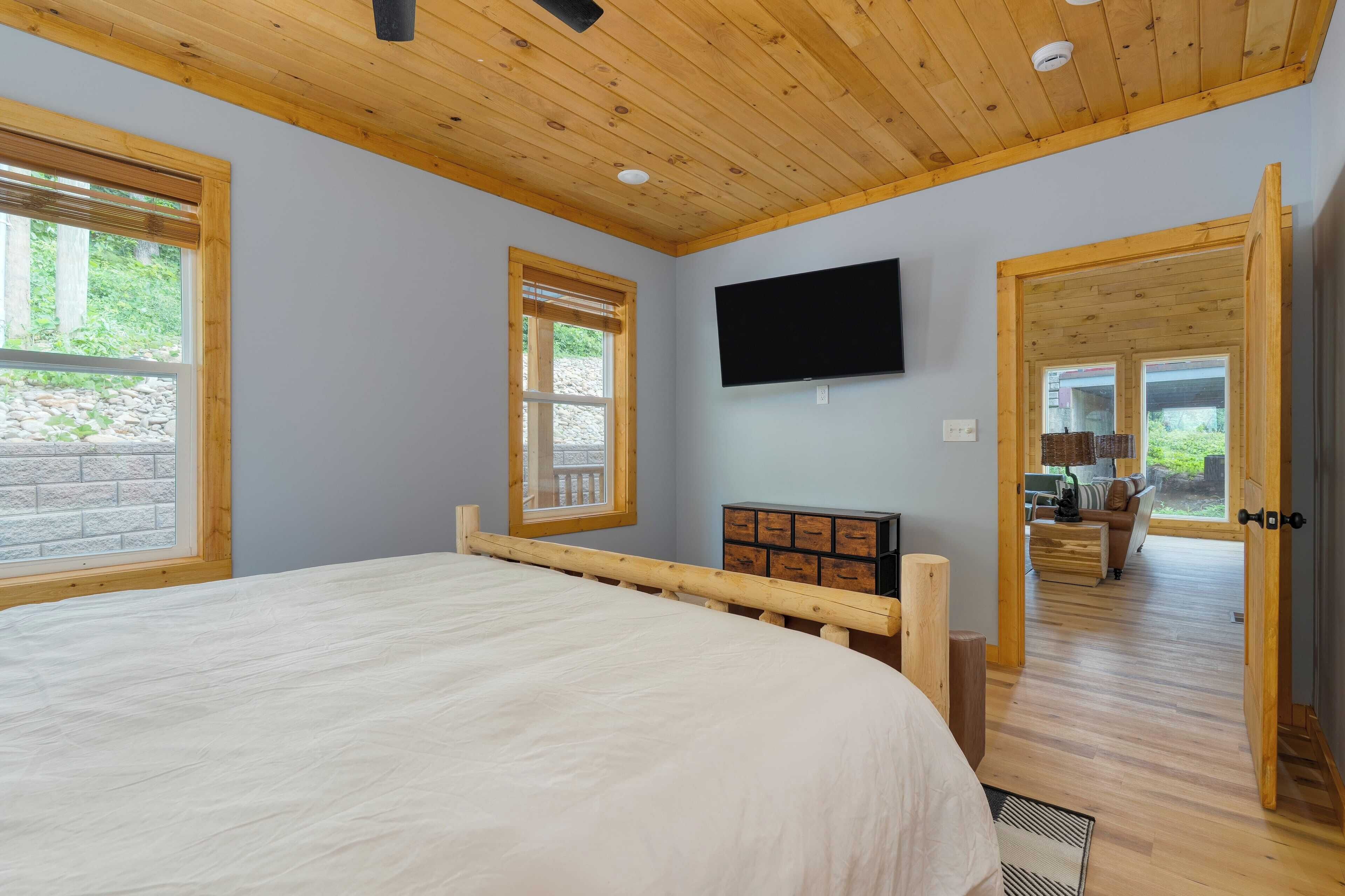 A bedroom with a large bed, wooden ceiling, a TV mounted on the wall, and a view of an outdoor seating area through an open door.