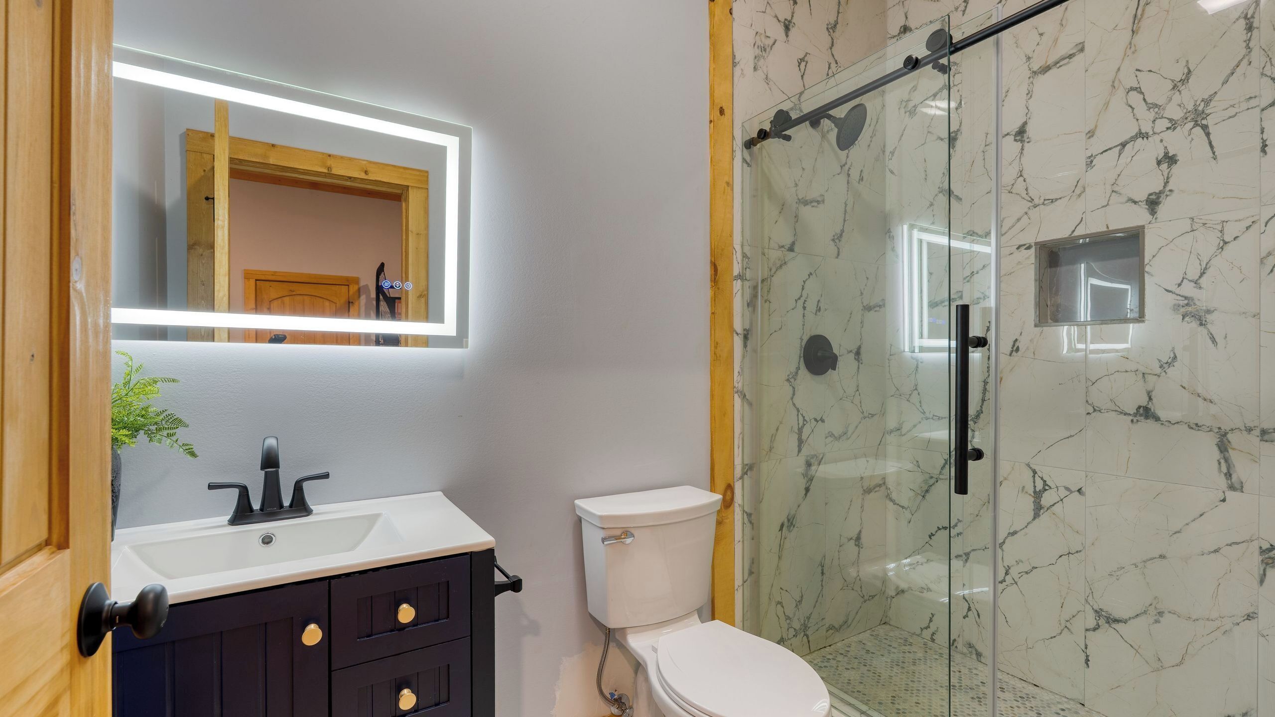 Un bagno moderno con un water bianco, un mobile lavabo scuro con pomelli dorati, un box doccia in vetro e uno schienale in marmo.