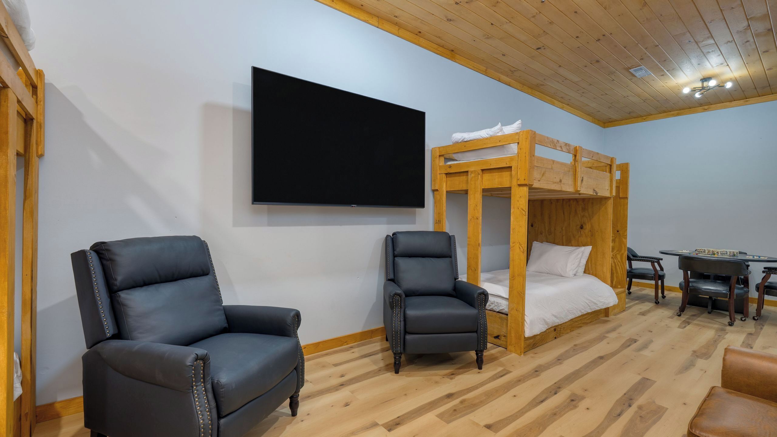 Quarto com cama de solteiro, duas poltronas de couro, televisão de tela plana e teto de madeira.