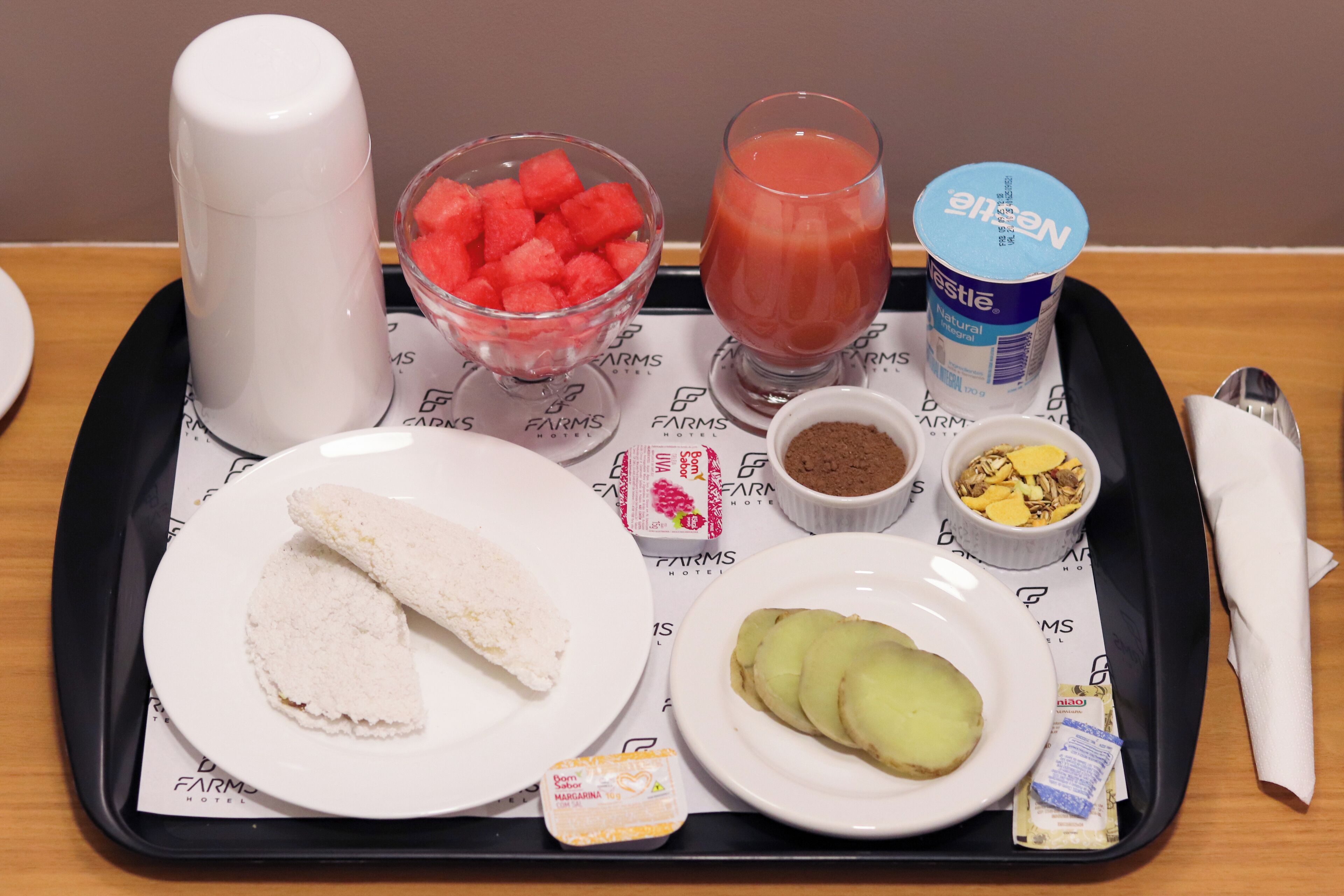 Een dienblad met eten, waaronder watermeloen, een glas sap, yoghurt en brood.