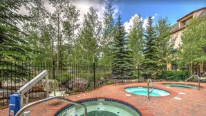 Un espace piscine doté de deux bains à remous, entouré d’une terrasse en briques, d’une clôture en métal et d’arbres en arrière-plan.