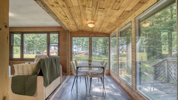 Ein gemütliches Zimmer mit Holzbalkendecke, großen Fenstern, einem Esstisch mit Stühlen und einer Couch mit gesteppter Decke.