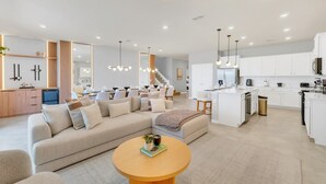 Una sala moderna con un amplio sofá seccional beige, una mesa de centro redonda de madera y una cocina con armarios blancos y una isla central.