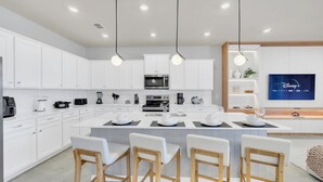 Una cocina moderna con armarios blancos, una isla central con taburetes y un televisor de pantalla plana montado en la pared.