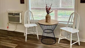 Een kamer met twee witte stoelen, een ronde tafel met een vaas, een raam met jaloezieën en een airconditioner.