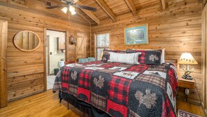 A wooden cabin bedroom with a bed, a ceiling fan, a nightstand with a lamp, and a painting on the wall. - Vista Cabin Getaway – Perfect Views + Dog Friendly (Blue Ridge)