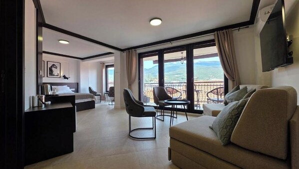 Habitación de hotel con cama, sofá, televisión, balcón con vistas y zona de comedor.