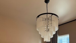 Ein Schlafzimmer mit am Wand montiertem Fernseher, einem Bett mit weißer Matratze, einem schwarzen Teddybären auf dem Bett, einer Hängelampe und einem Fenster mit Vorhängen.