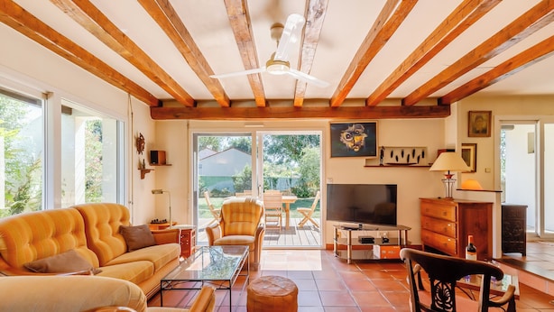 Ein geräumiges Wohnzimmer mit einer Couch, Sesseln, einem Couchtisch und einem Fernseher. Die Holzbalkendecke ist sichtbar und eine Glastür öffnet sich zu einem Außenbereich.