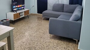 Una sala de estar con un sofá gris, un televisor sobre un soporte blanco, una mesita y una ventana con persianas.