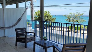 A covered outdoor area with a hammock, two chairs, a small table, and a view of the sea.