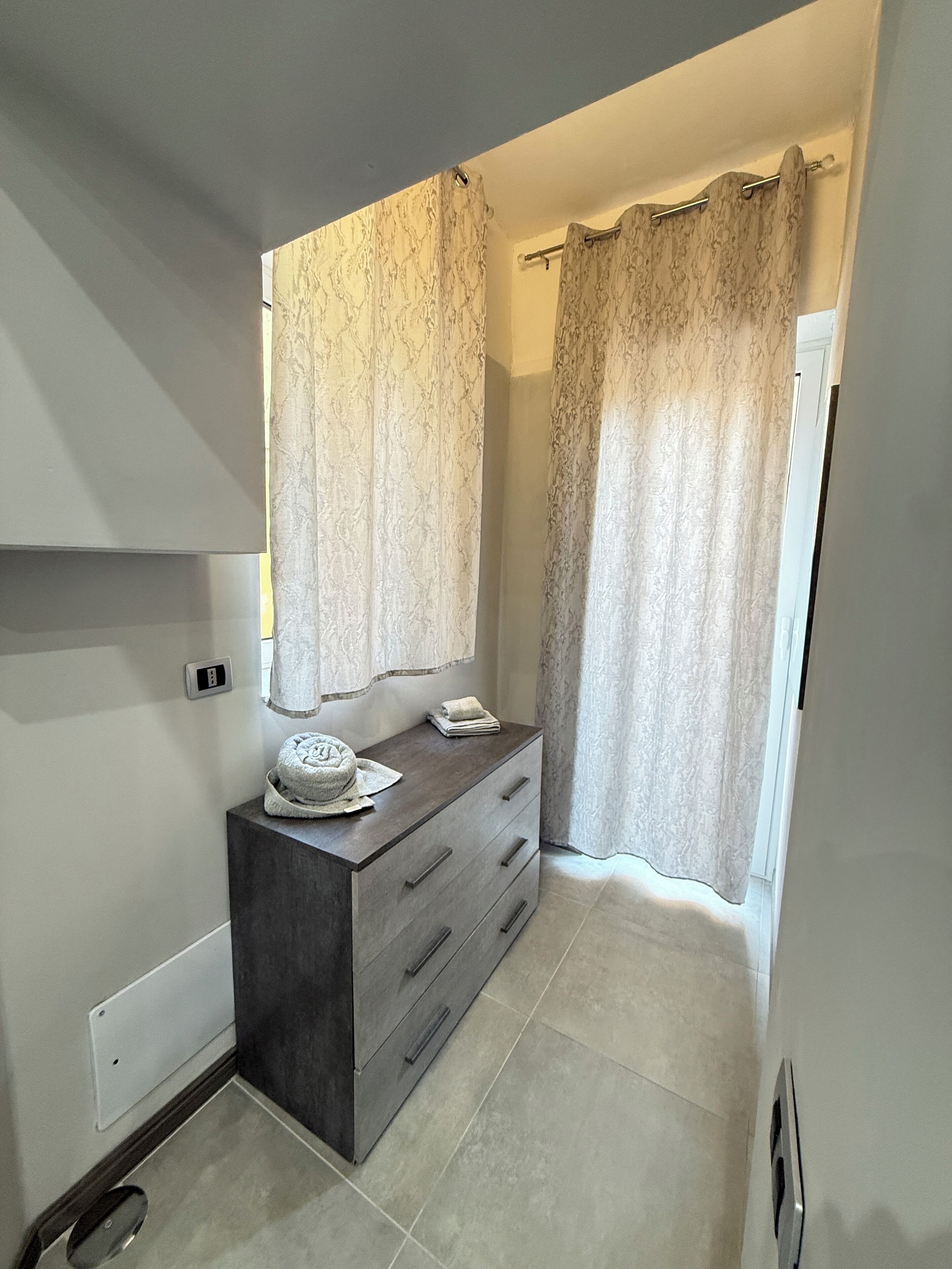 A bathroom with a dark wooden vanity, a towel, and a hat on it. There is a light switch on the wall and a curtain on the window.