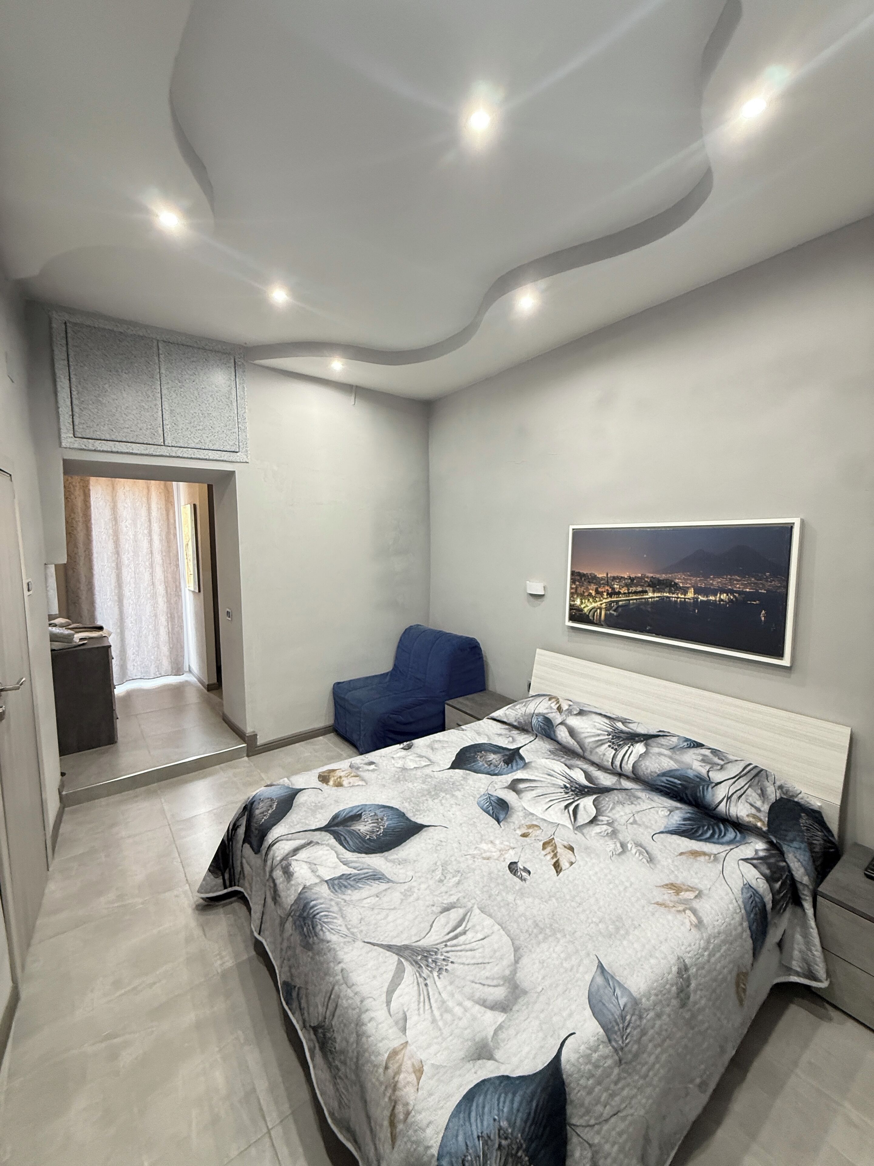 A bedroom with a bed, a chair, a painting, and a view of a cityscape.
