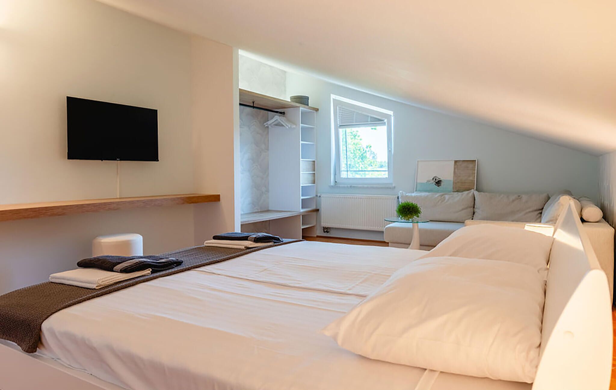 Ein modernes Schlafzimmer mit einem großen Bett, einem an der Wand montierten Fernseher, einem begehbaren Kleiderschrank und einem Fenster, das für natürliches Licht sorgt.