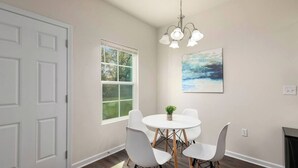 A dining area with a round table, four white chairs, a small plant, and a painting on the wall. - Southern Gem, Game Rm, Fire Pit, Near Coliseum (Greensboro)
