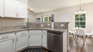 A modern kitchen with white cabinets, granite countertops, and a dishwasher. There is a dining area with a table and chairs. - Southern Gem, Game Rm, Fire Pit, Near Coliseum (Greensboro)