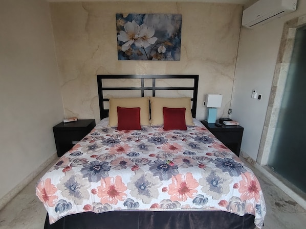 Ein Bett mit floraler Bettdecke, zwei Nachttische mit Lampen, eine Wand-Klimaanlage und ein Blumenbild über dem Bett.