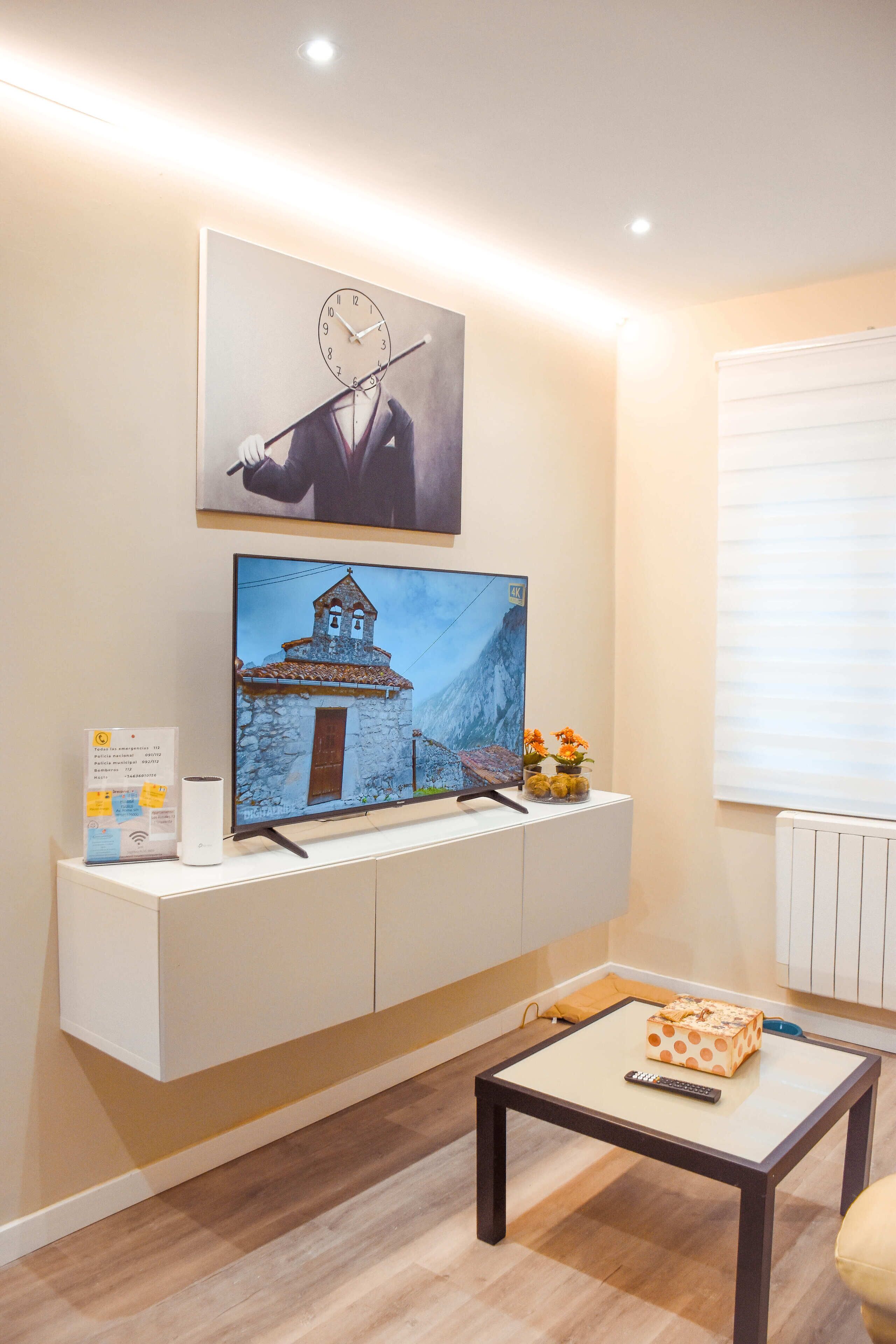 A room with a mounted television displaying a scenic image, a white console table with a remote control, a small vase with flowers, and a window with blinds.