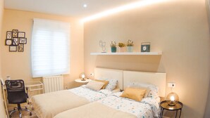 A bedroom with two beds, a bedside table, a chair, a radiator, and a shelf with decorative items. - Perfecta UbicaciÓn Para Conocer Asturias (Oviedo)