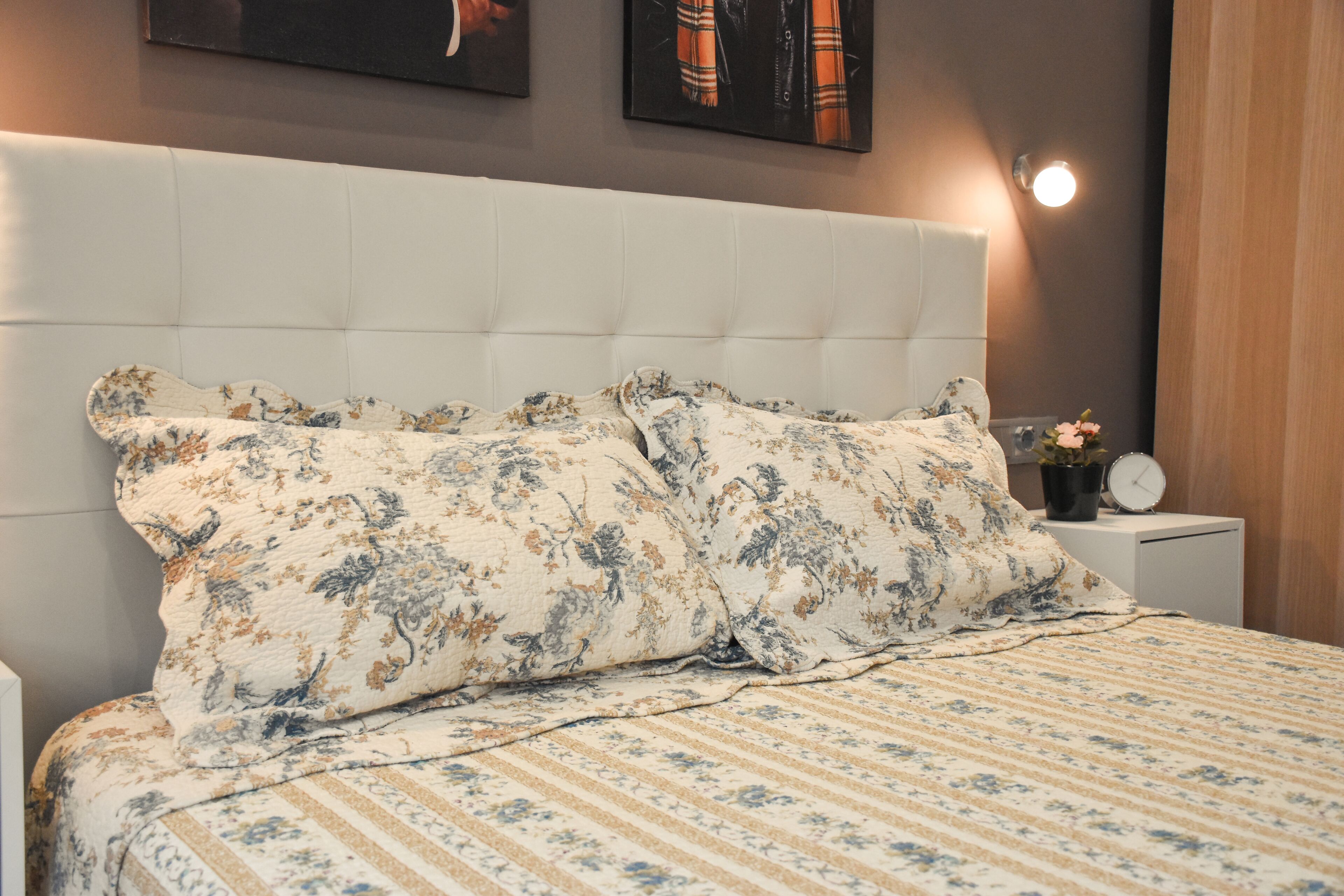 A neatly made bed with floral bedding, a white headboard, and a bedside table with a clock and a small plant.