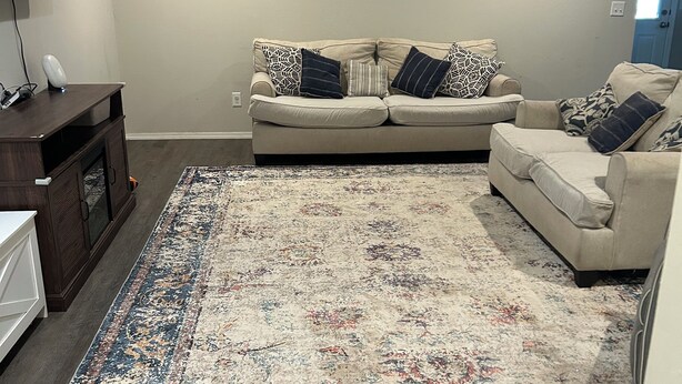 A living room with a beige sofa, patterned cushions, a large rug, and a wall-mounted TV.