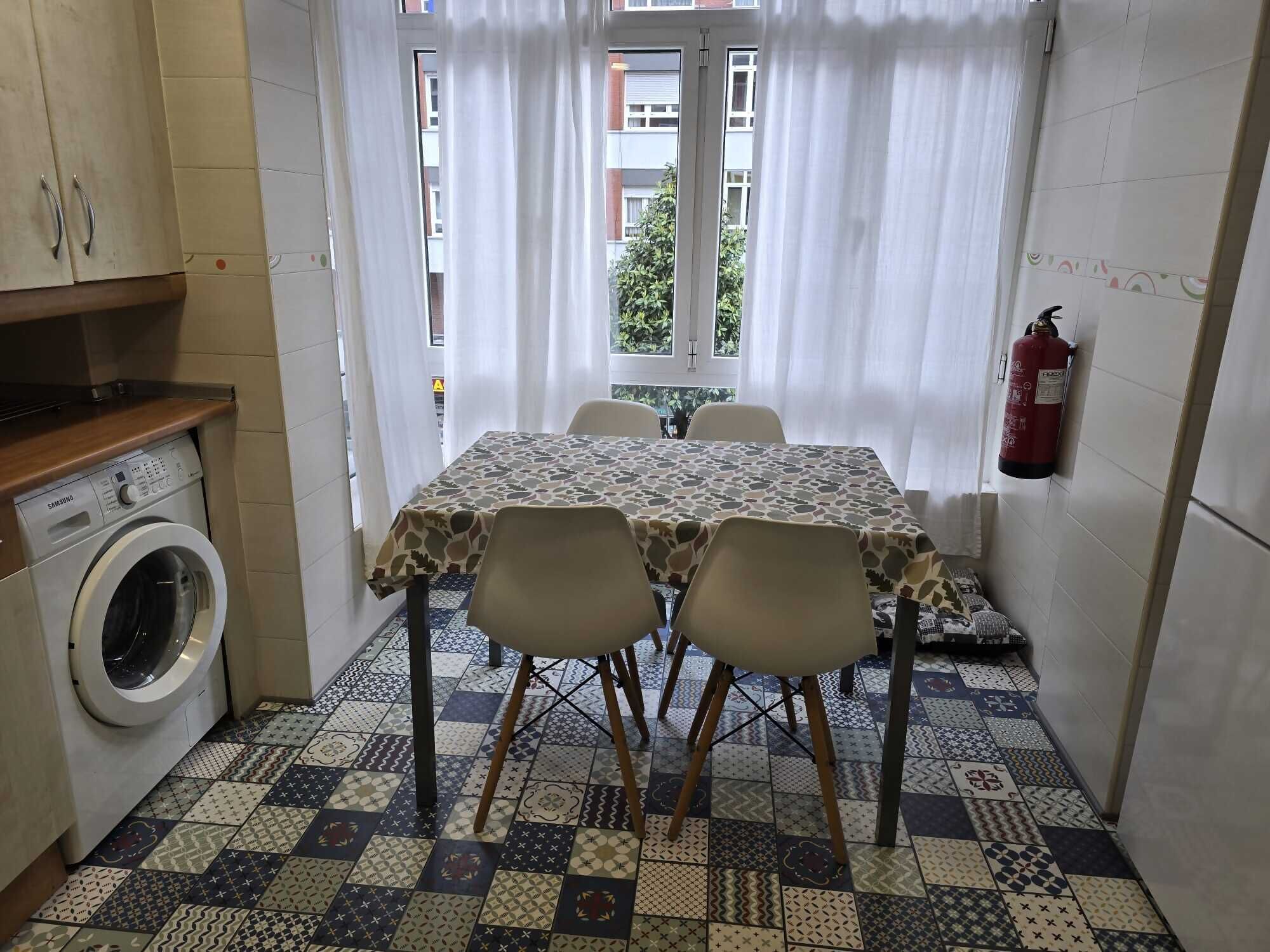 Een kleine keuken met een wasmachine, een tafel met stoelen en een brandblusser aan de muur.