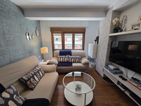 Un salon comprenant un canapé, une table basse, un téléviseur et une fenêtre donnant sur des immeubles.