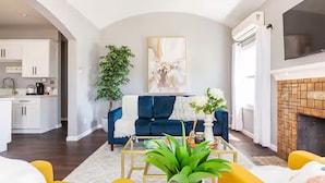 A modern living room with a blue sofa, a fireplace, a kitchen in the background, and a large plant in the center.