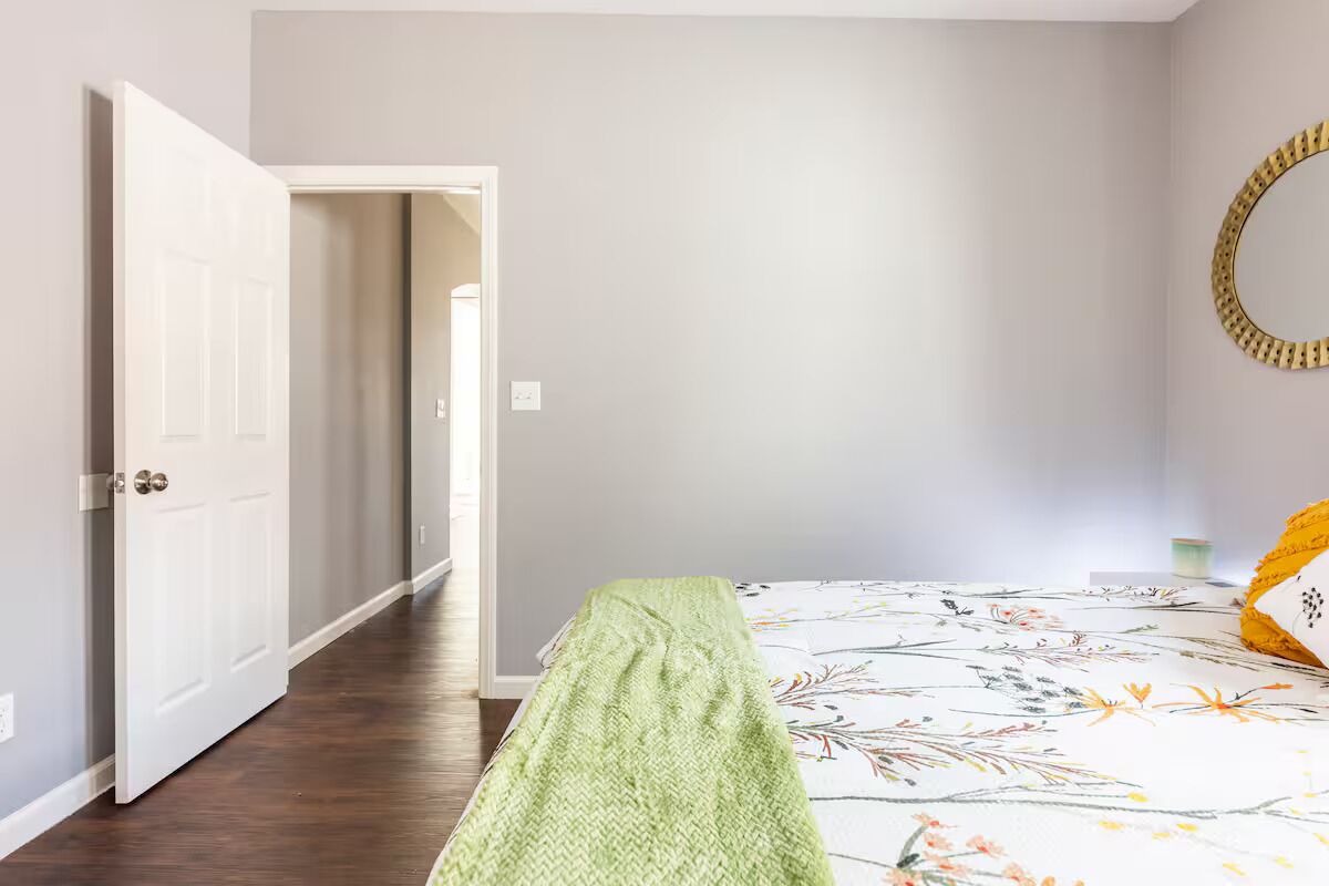 A bedroom with a bed, a green throw blanket, a floral bedspread, a round mirror, and a white door leading to another room.