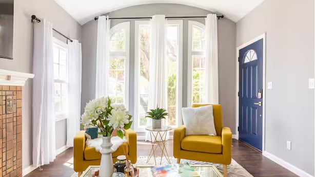 A living room with a fireplace, two yellow armchairs, a coffee table, and a large window with curtains.