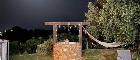 A stone fire pit with a wooden pergola overhead, surrounded by grass and a hammock.