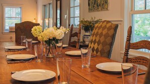 A dining room with a wooden table set for six, two hanging pendant lights, and a large window with a view of greenery.