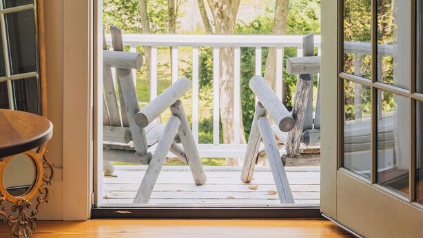 A room with a wooden floor, a mirror, a green wall hanging, and a view of a wooden deck with chairs and trees.