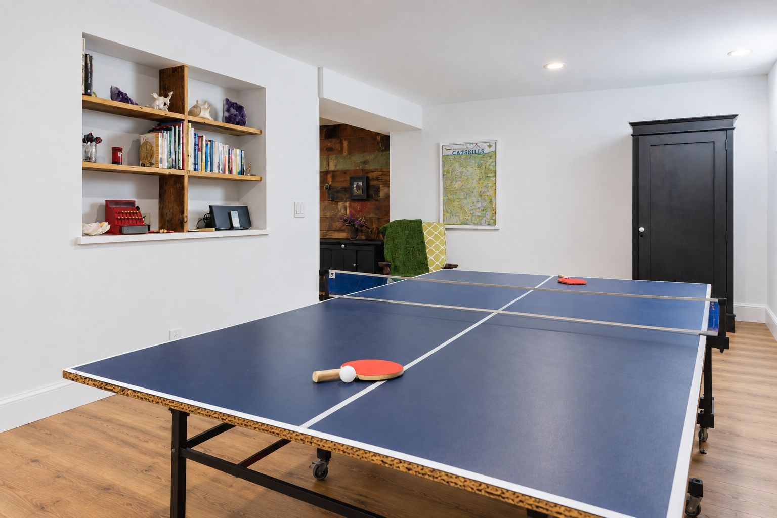 Une table de ping-pong avec deux raquettes, une bibliothèque remplie de livres et d’objets décoratifs, et une carte accrochée au mur.