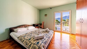 A bedroom with a bed, a chair, a desk, and a wardrobe. There is a view of mountains and trees through a glass door.