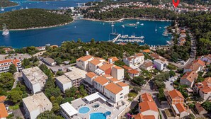 An aerial view of a coastal town with a marina, buildings with red-tiled roofs, and a large swimming pool with sun loungers.