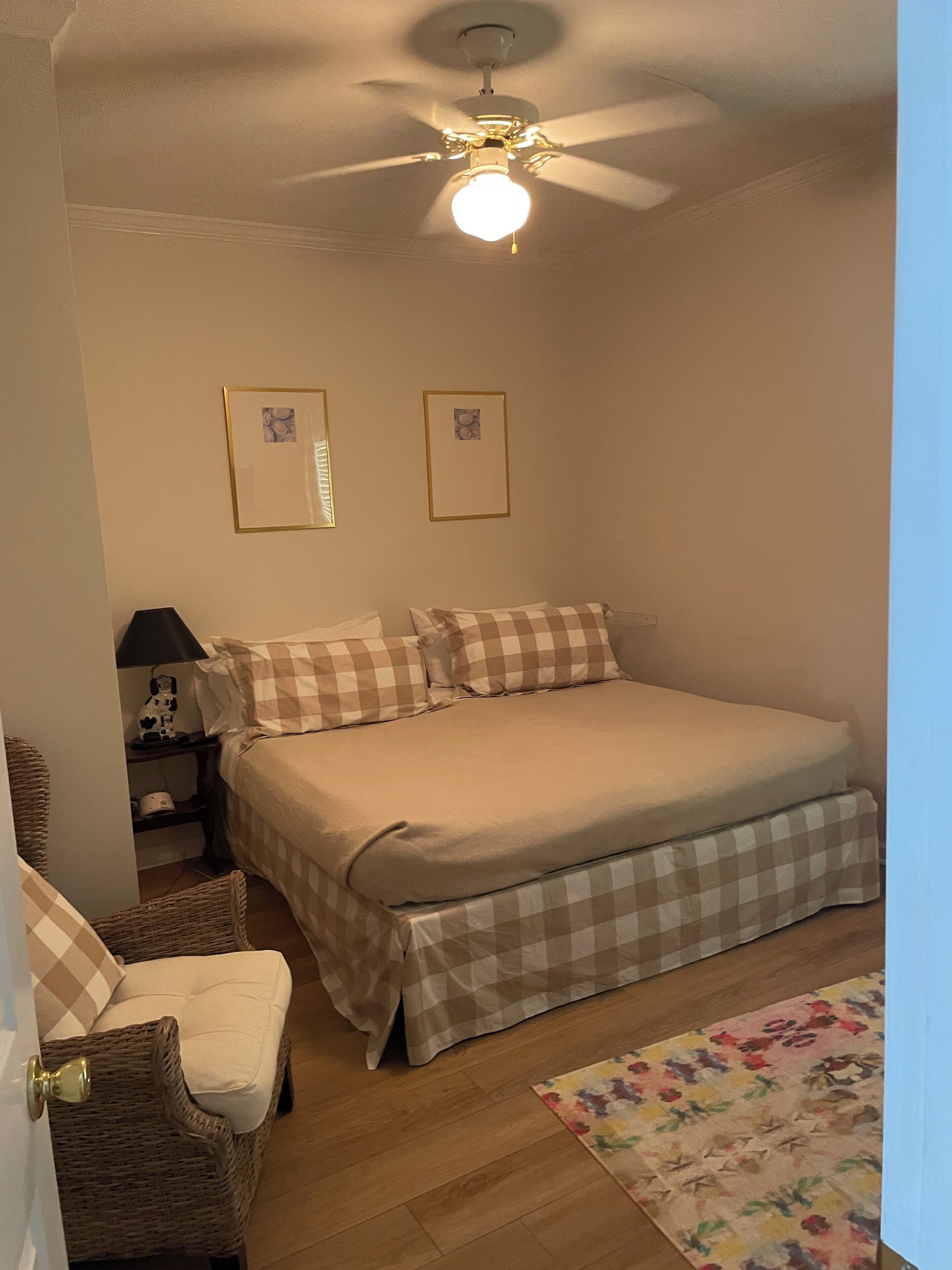 Een slaapkamer met een bed, een plafondventilator, twee ingelijste foto's, een nachtkastje met een lamp, een rieten stoel en een bloemenkleed.