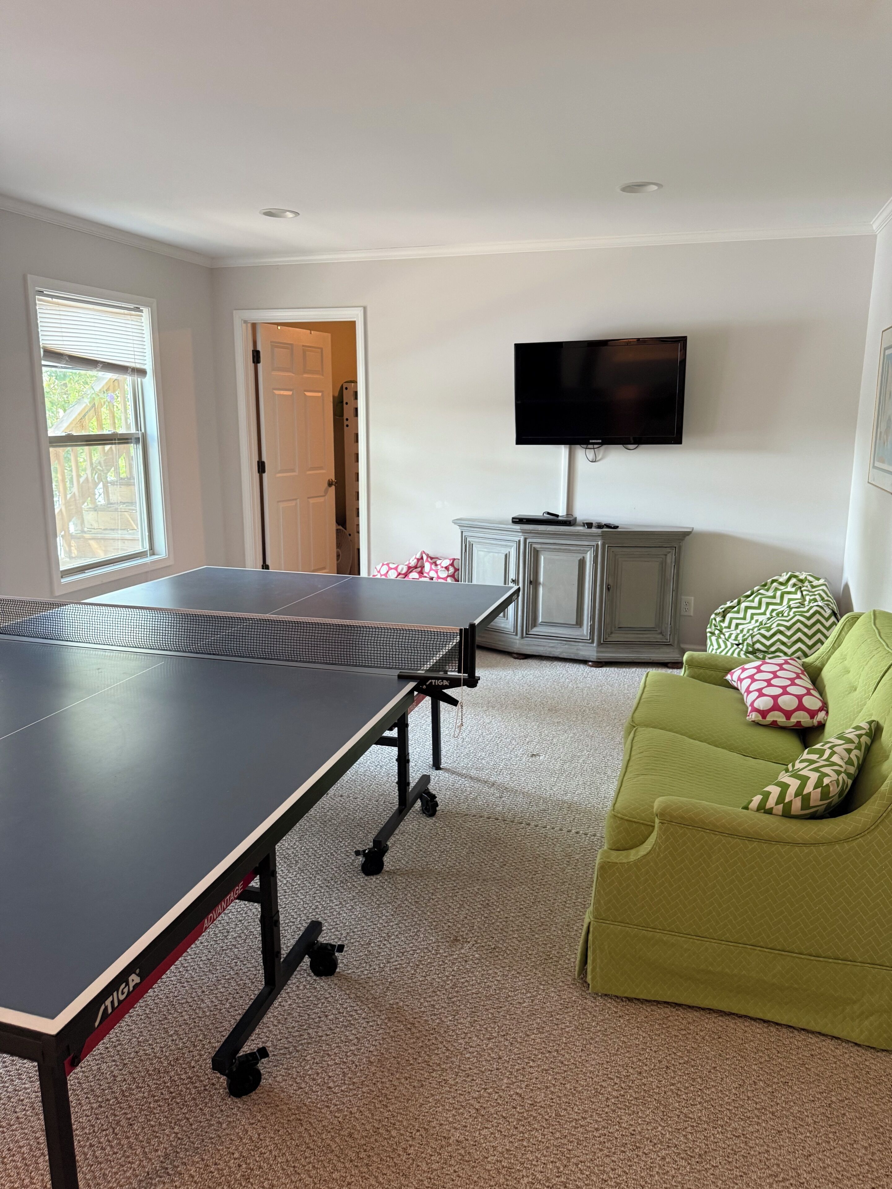 Une pièce avec une table de ping-pong, un téléviseur, un canapé vert et une porte qui donne sur une autre pièce.