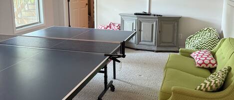 Una habitación con mesa de ping pong, televisión, sofá verde y puerta que da a otra estancia.
