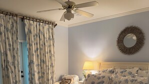 A bedroom with a bed, a ceiling fan, a bedside lamp, and a decorative mirror on the wall.