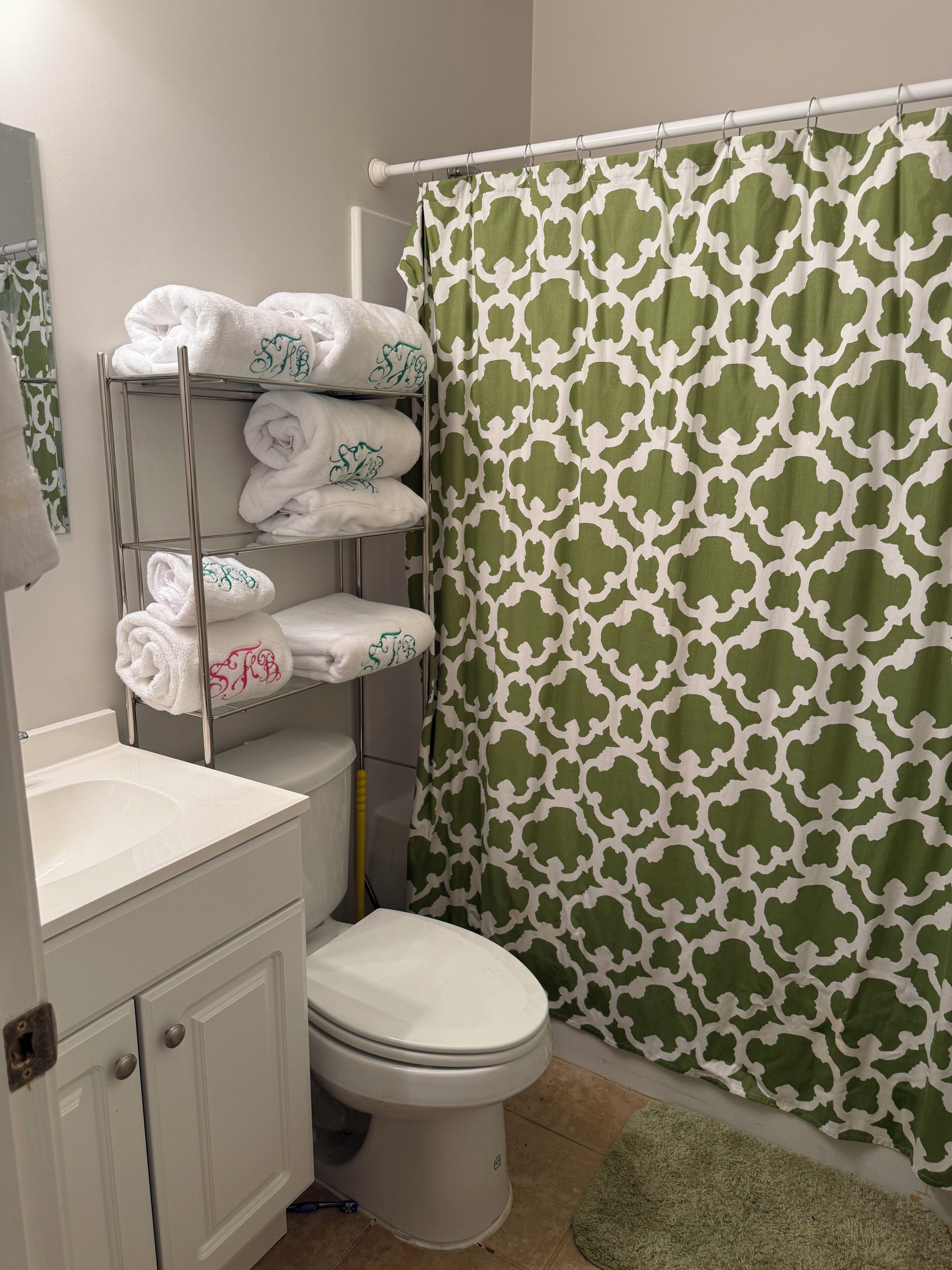Un baño con inodoro, lavamanos, toallero con toallas dobladas con cuidado y cortina de ducha estampada en verde y blanco.